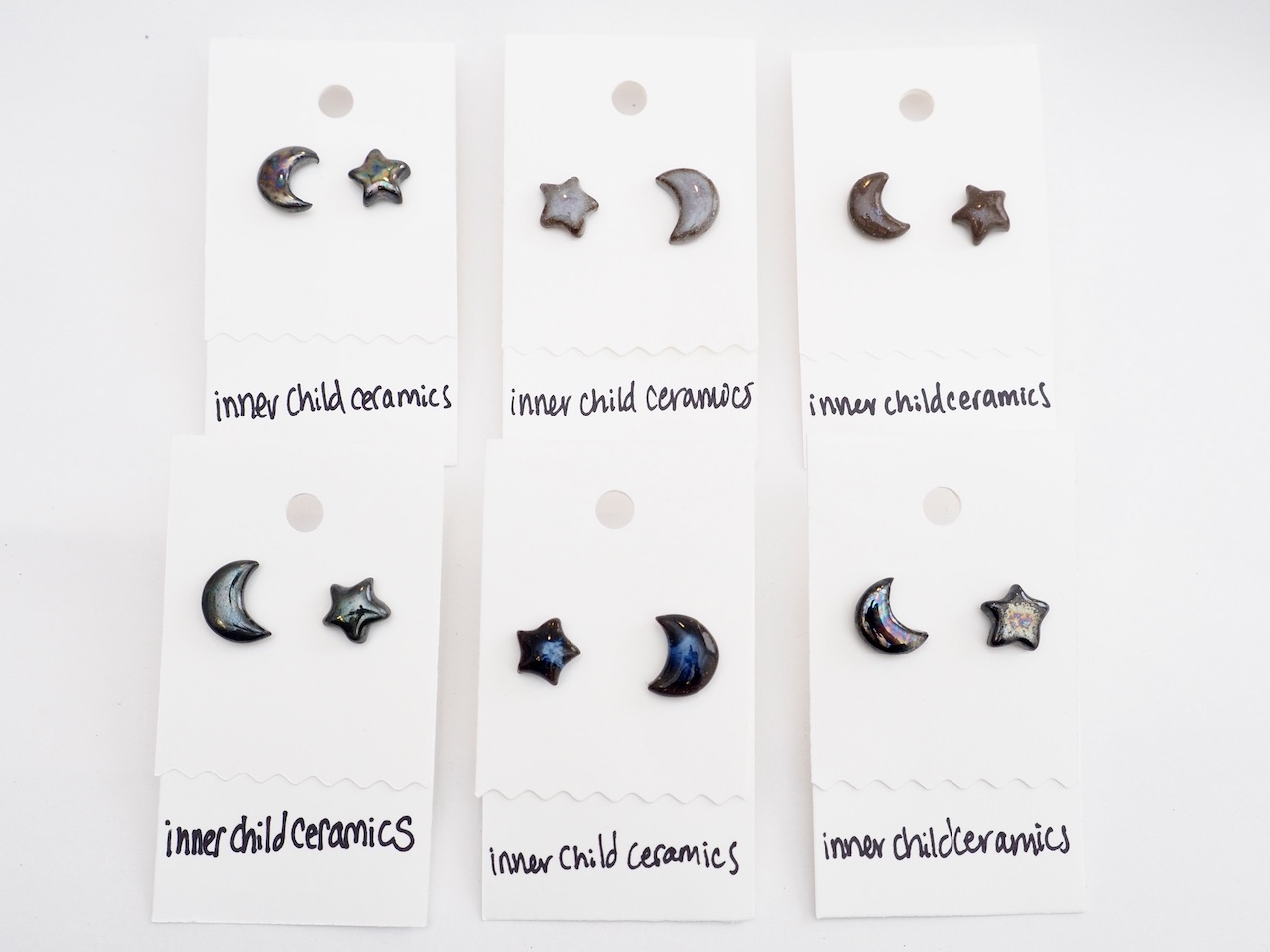 Star & Moon Earrings - 6 Pairs A grid arrangement of six pairs of handmade ceramic star and moon stud earrings on white display cards. The collection showcases various glaze finishes, including iridescent "oilslick," deep "Midnight Indigo" blue swirls, and speckled neutral tones. Each card has the handwritten "inner child ceramics" branding at the bottom, highlighting the unique, handcrafted variation across the entire celestial collection.