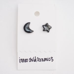 A pair of mismatched ceramic stud earrings on a white card, consisting of one crescent moon and one five-pointed star. These earrings feature a "Black and Emerald" finish, a deep, dark green with a subtle metallic or speckled sheen. The brand name "inner child ceramics" is handwritten in black at the bottom of the display card.