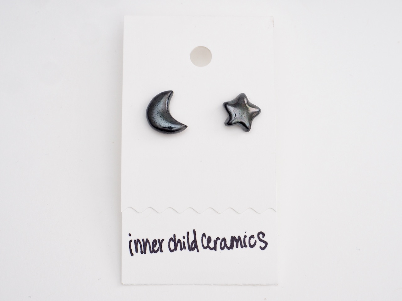 Black Emerald Star & Moon Earrings A pair of mismatched ceramic stud earrings on a white card, consisting of one crescent moon and one five-pointed star. These earrings feature a "Black and Emerald" finish, a deep, dark green with a subtle metallic or speckled sheen. The brand name "inner child ceramics" is handwritten in black at the bottom of the display card.