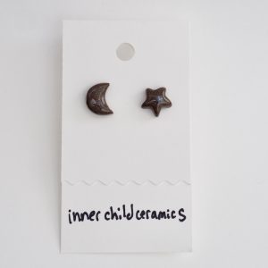 A pair of mismatched ceramic stud earrings—one star and one crescent moon—on a white display card. These earrings feature a "Chocolate" finish, characterized by a deep, glossy brown color with fine, sparkling speckles throughout. The brand name "inner child ceramics" is handwritten in black at the bottom.