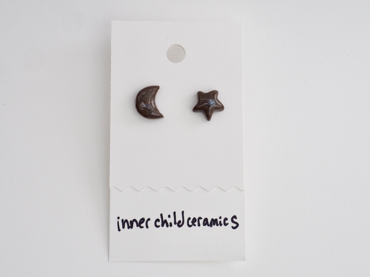 Chocolate Star & Moon A pair of mismatched ceramic stud earrings—one star and one crescent moon—on a white display card. These earrings feature a "Chocolate" finish, characterized by a deep, glossy brown color with fine, sparkling speckles throughout. The brand name "inner child ceramics" is handwritten in black at the bottom.