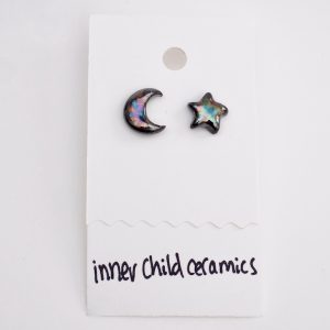 A pair of mismatched ceramic stud earrings on a white card. One earring is shaped like a crescent moon and the other like a five-pointed star. Both feature a dark, glossy "oilslick" finish with iridescent swirls of blue, purple, and gold. The white display card has the brand name "inner child ceramics" handwritten in black ink at the bottom.