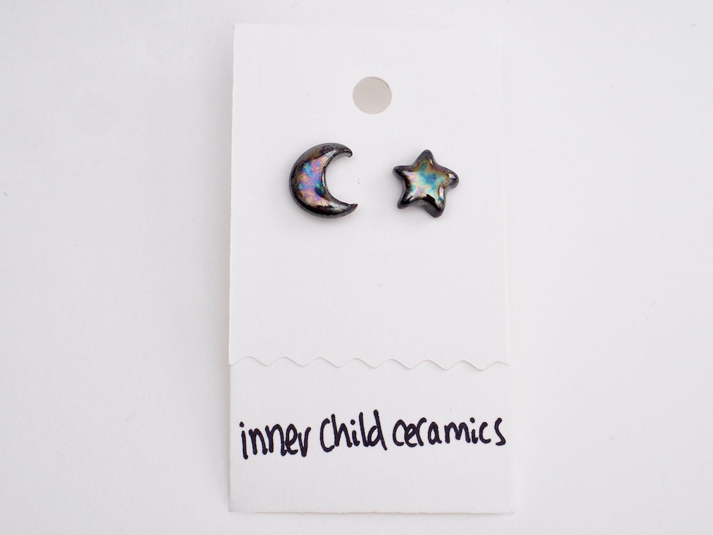 Cosmic Oil Slick Star & Moon A pair of mismatched ceramic stud earrings on a white card. One earring is shaped like a crescent moon and the other like a five-pointed star. Both feature a dark, glossy "oilslick" finish with iridescent swirls of blue, purple, and gold. The white display card has the brand name "inner child ceramics" handwritten in black ink at the bottom.
