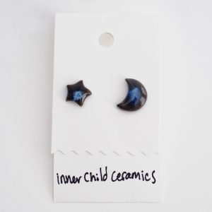 A single pair of mismatched ceramic stud earrings (one star and one crescent moon) on a white display card. These earrings feature a "Midnight Indigo" finish, characterized by a dark black base with a luminous, swirling deep blue center that resembles a nebula or a night sky. The card is labeled with the handwritten text "inner child ceramics" at the bottom.