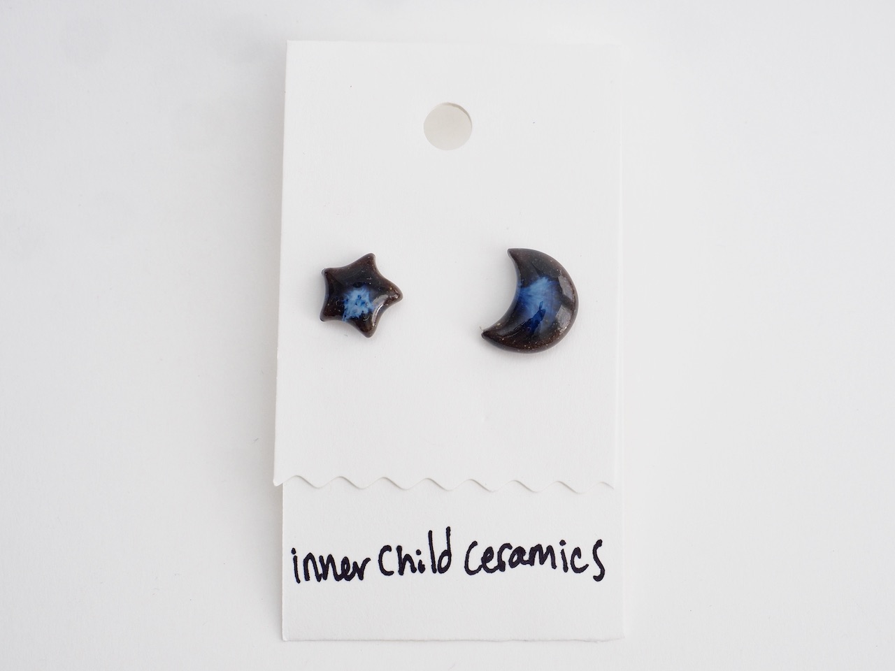 Midnight Indigo Star & Moon A single pair of mismatched ceramic stud earrings (one star and one crescent moon) on a white display card. These earrings feature a "Midnight Indigo" finish, characterized by a dark black base with a luminous, swirling deep blue center that resembles a nebula or a night sky. The card is labeled with the handwritten text "inner child ceramics" at the bottom.