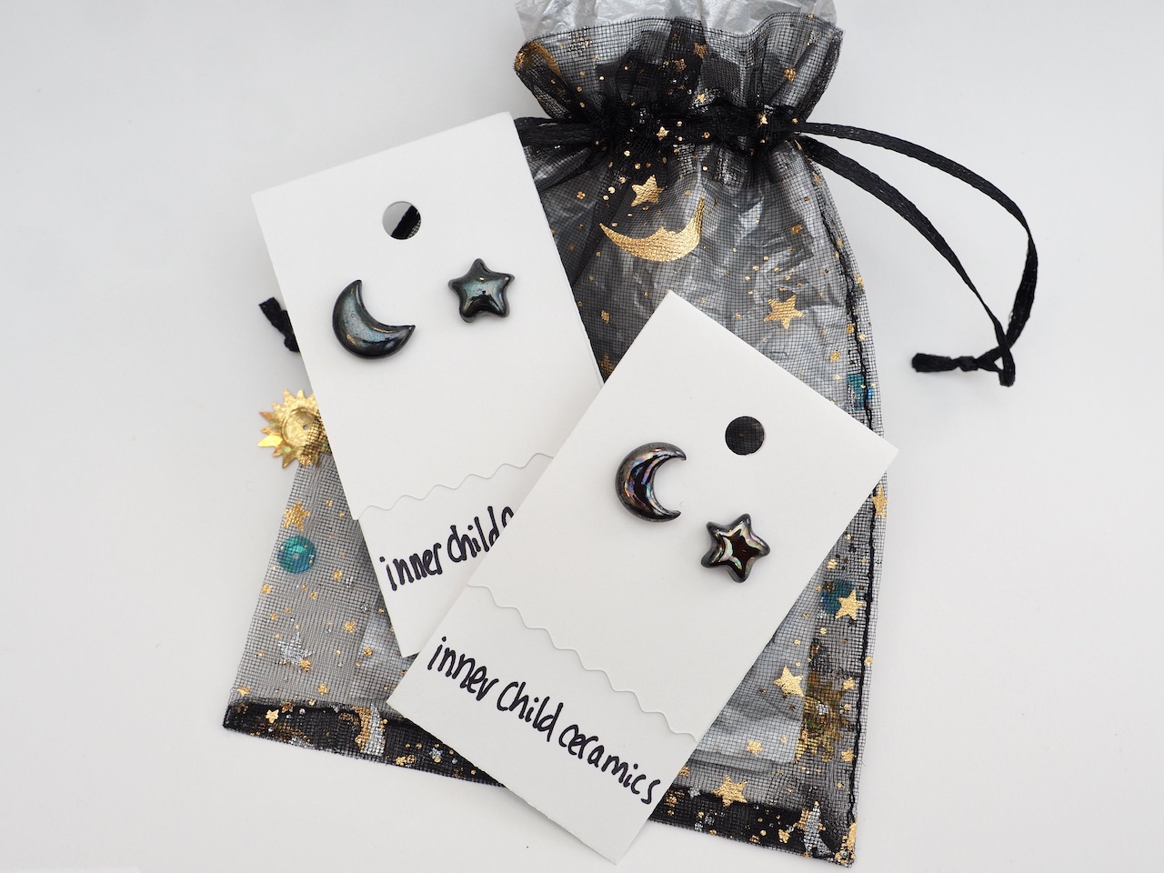 Black Emerald and Cosmic Oil Slick Star & Moon Earrings Paired Up Two pairs of mismatched ceramic stud earrings by Inner Child Ceramics, displayed on white cards over a black organza gift bag. Each set includes a crescent moon and a five-pointed star earring with a glossy, dark "oilslick" iridescent glaze. The sheer black drawstring bag is decorated with gold-foiled stars and moons, mirroring the celestial design of the jewelry. The brand name "inner child ceramics" is handwritten in black at the bottom of the white display cards.