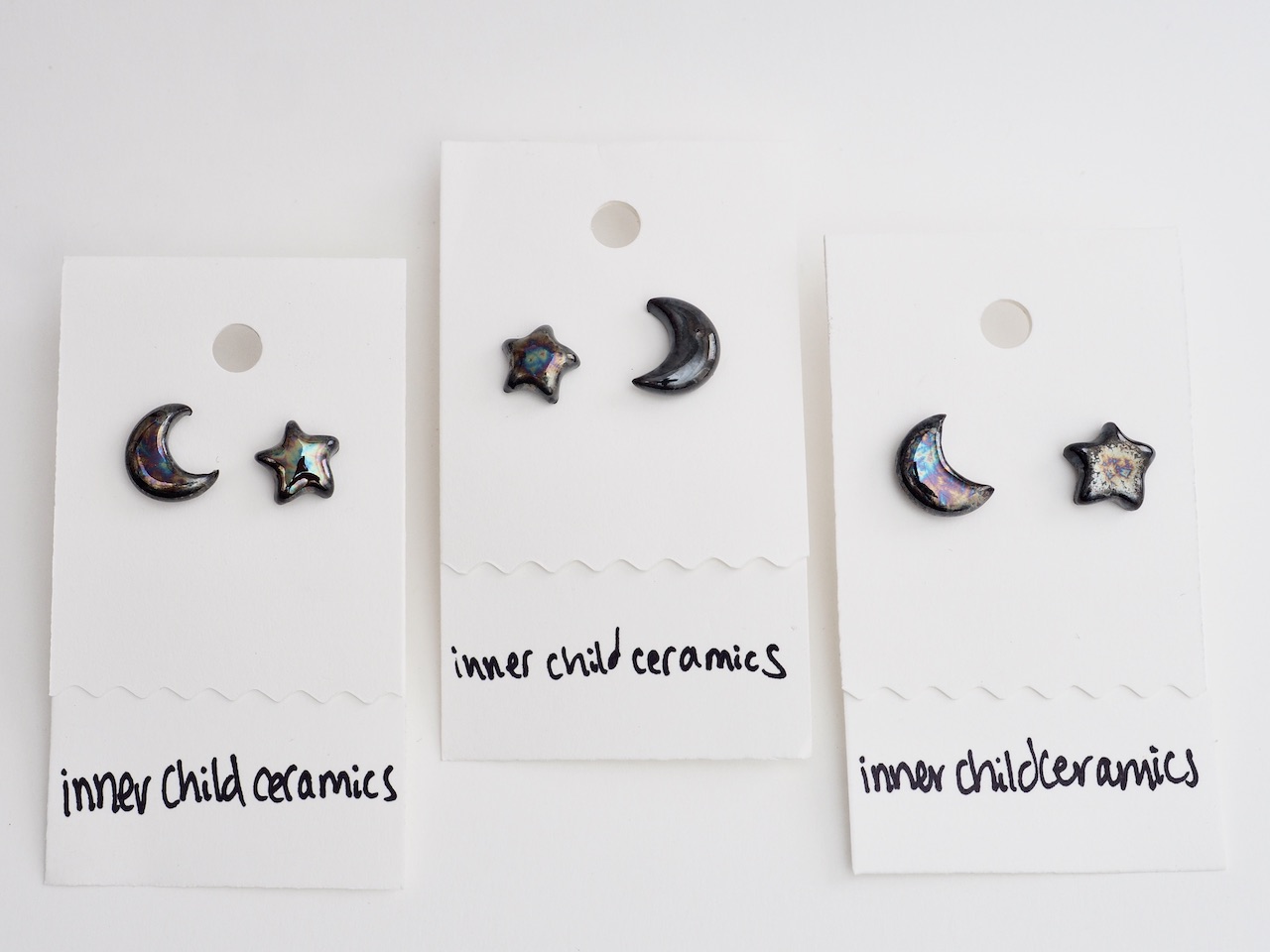 Trio of Cosmic Oil Slick Star & Moons A horizontal arrangement of three pairs of handmade ceramic stud earrings. Each pair consists of one crescent moon and one star earring, all finished with a high-shine, iridescent glaze that reflects blue and purple tones. They are mounted on white display cards that feature the handwritten text "inner child ceramics" at the bottom.