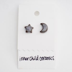 A pair of mismatched ceramic stud earrings—one star and one crescent moon—on a white display card. These earrings feature a pale, speckled finish that appears as a soft off-white or light lavender with dark, organic edges and a high-gloss sheen. The card is labeled with the handwritten text "inner child ceramics" at the bottom.